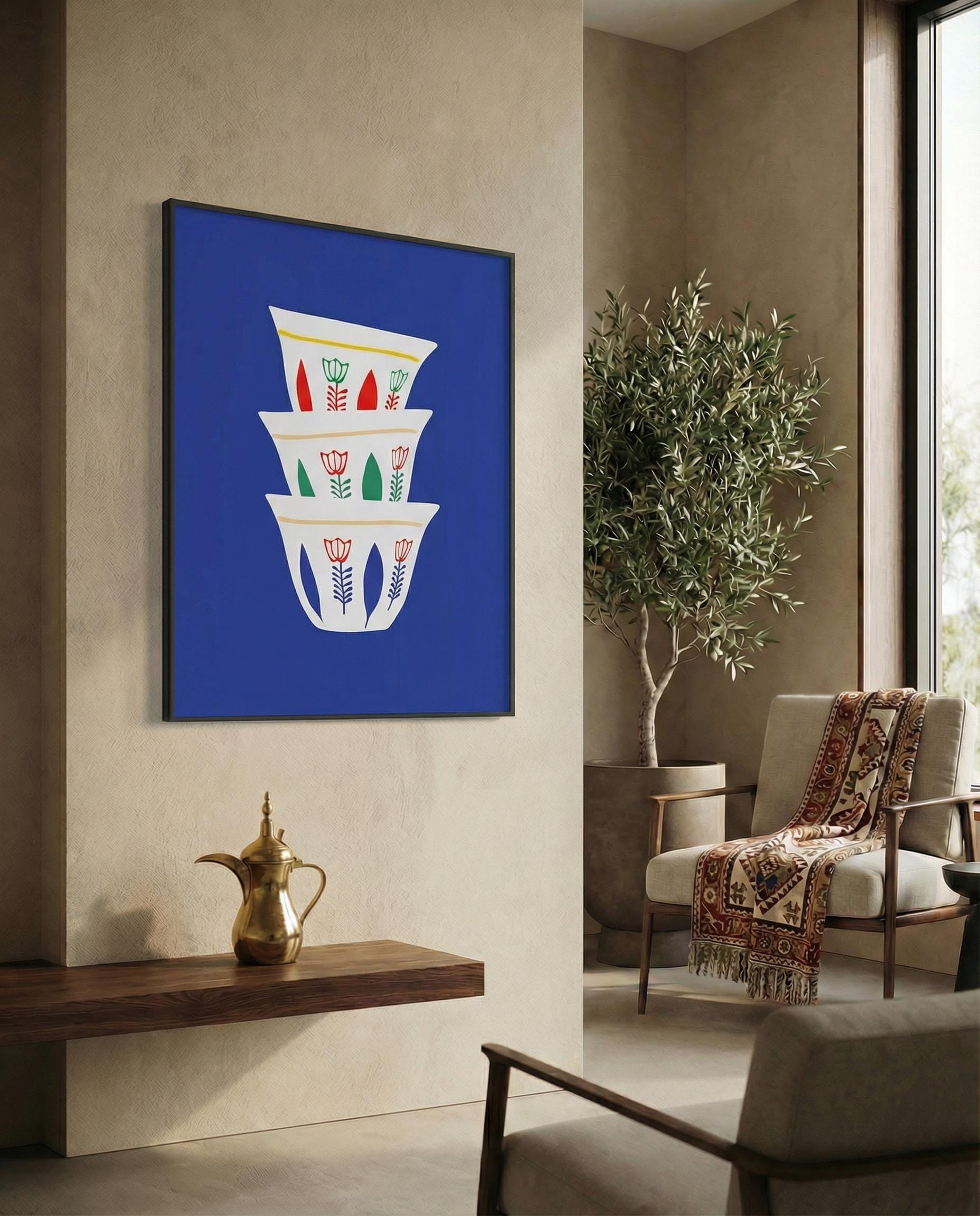 A framed art print featuring a stack of three white coffee cups with floral designs in red, green, and yellow against a vibrant blue background. The artwork is displayed in a modern living space.