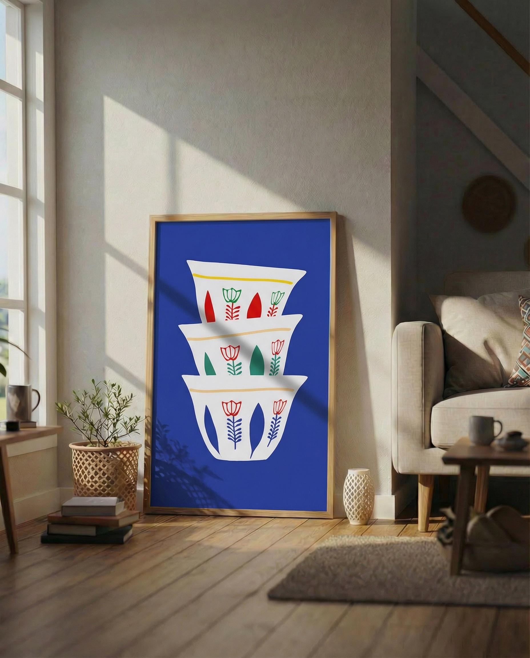 A framed art print featuring a stack of white coffee cups with floral designs against a vibrant blue background. The cups have red, green, and yellow accents. The artwork is displayed in a living room setting with natural light.