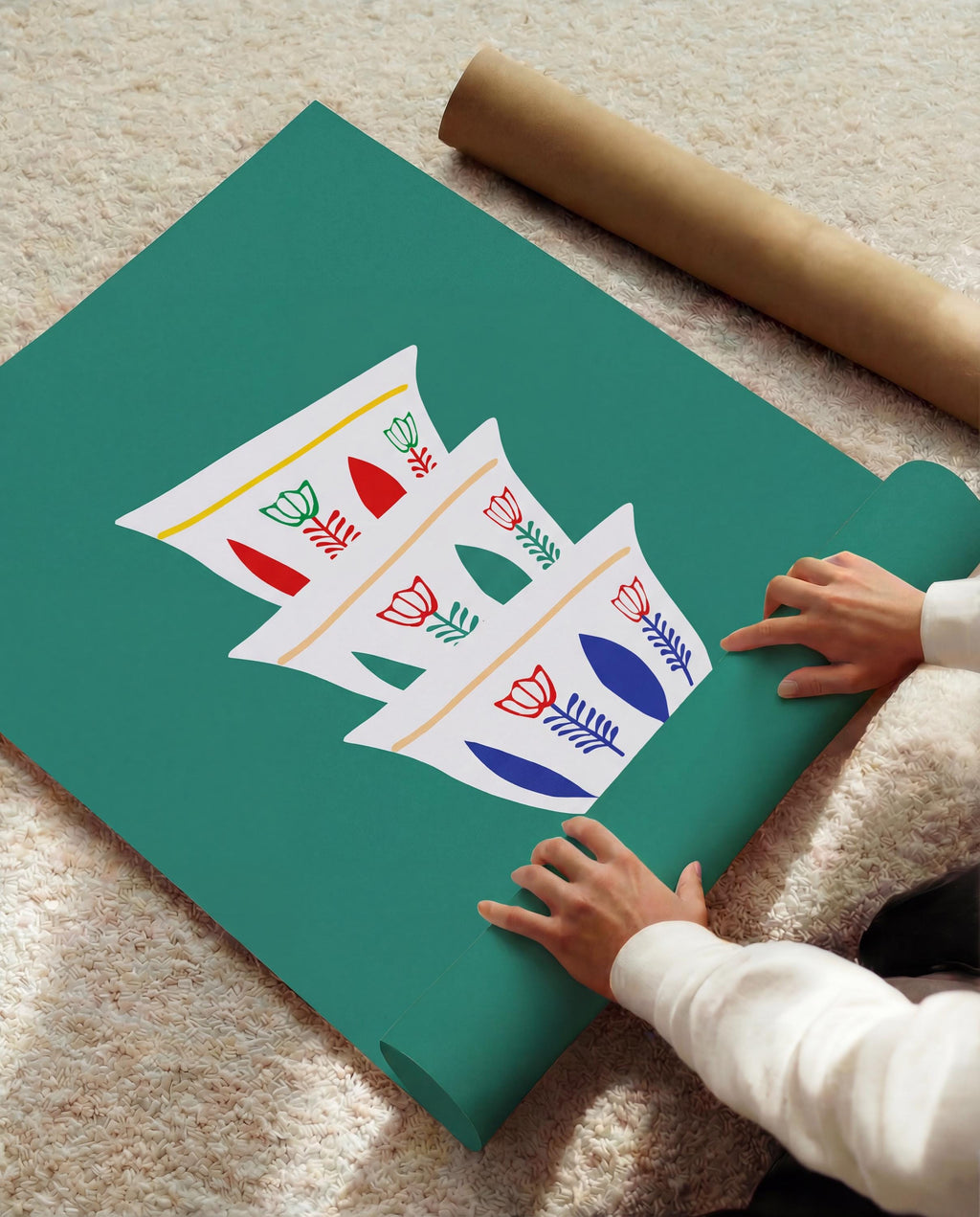 A teal-colored poster featuring a stack of three white coffee cups with floral designs in red, green, and blue. The poster is partially rolled out, with a brown paper tube visible in the background. The image is set on a light-colored rug.