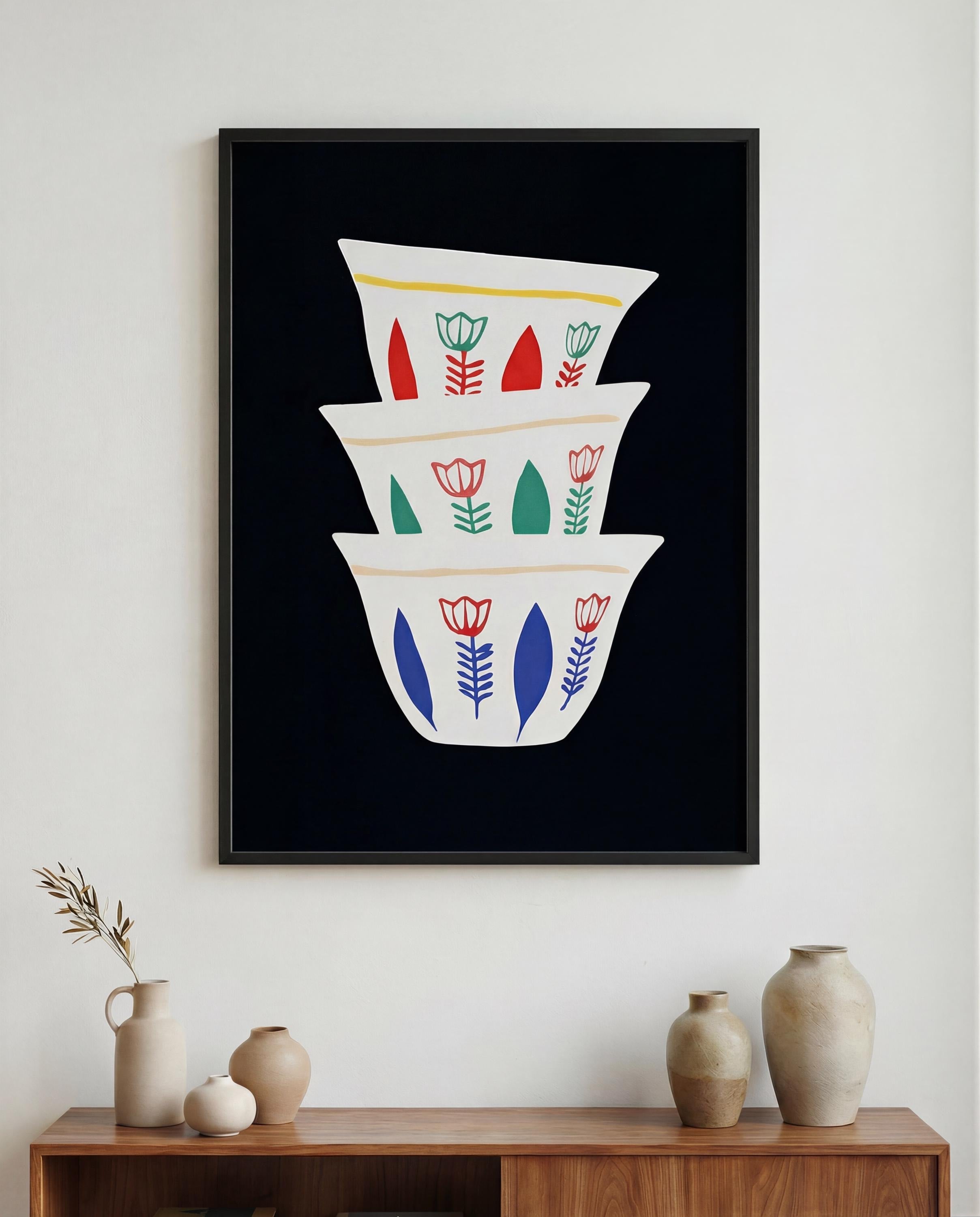 A framed art print featuring a stack of three white bowls with floral designs against a black background. The bowls have red, green, and blue floral patterns. The artwork is displayed above a wooden cabinet with vases.