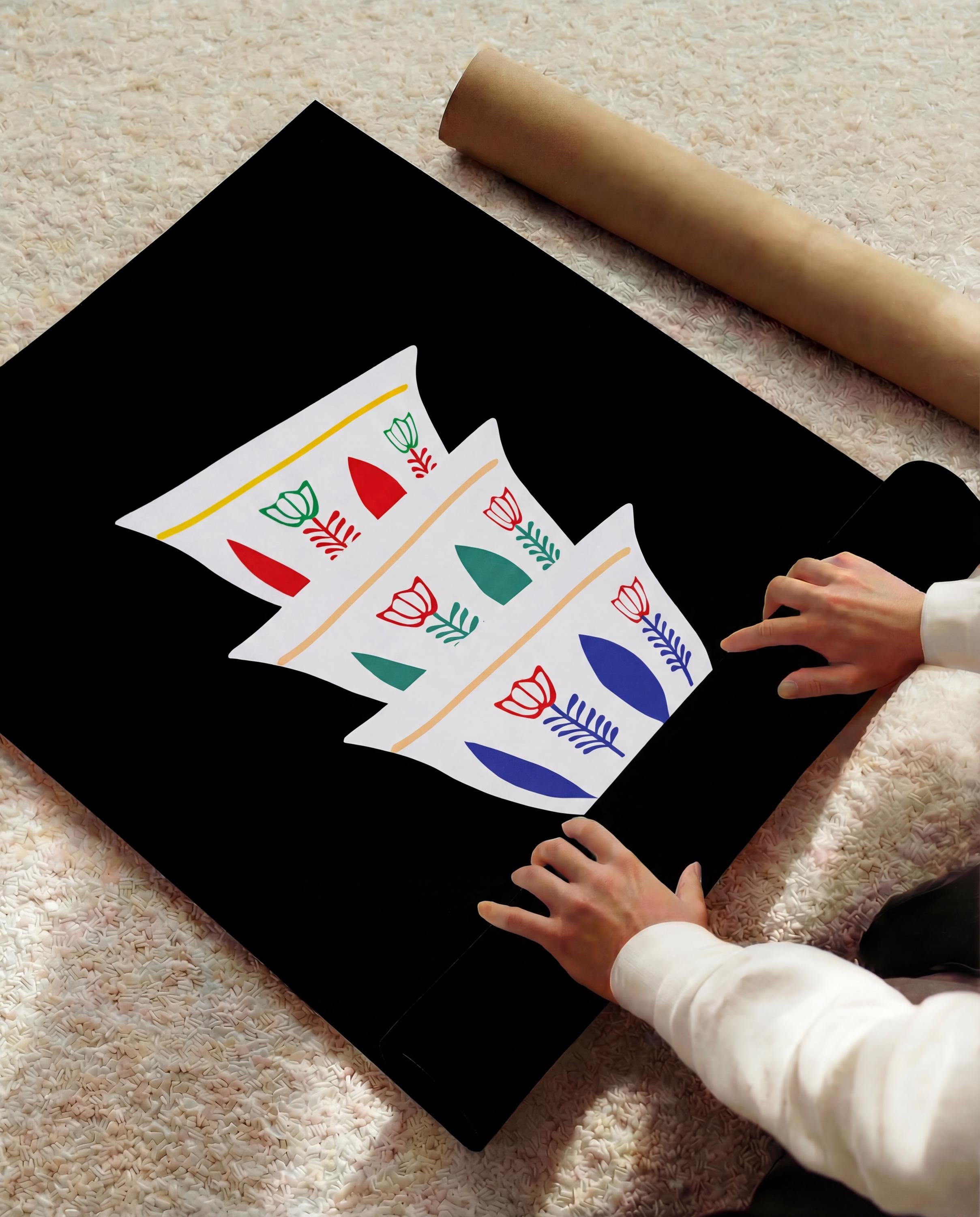 A black poster with a white design of three stylized coffee cups, each with colorful floral and leaf patterns. A rolled-up brown paper tube rests nearby. The poster is being unrolled by a person.