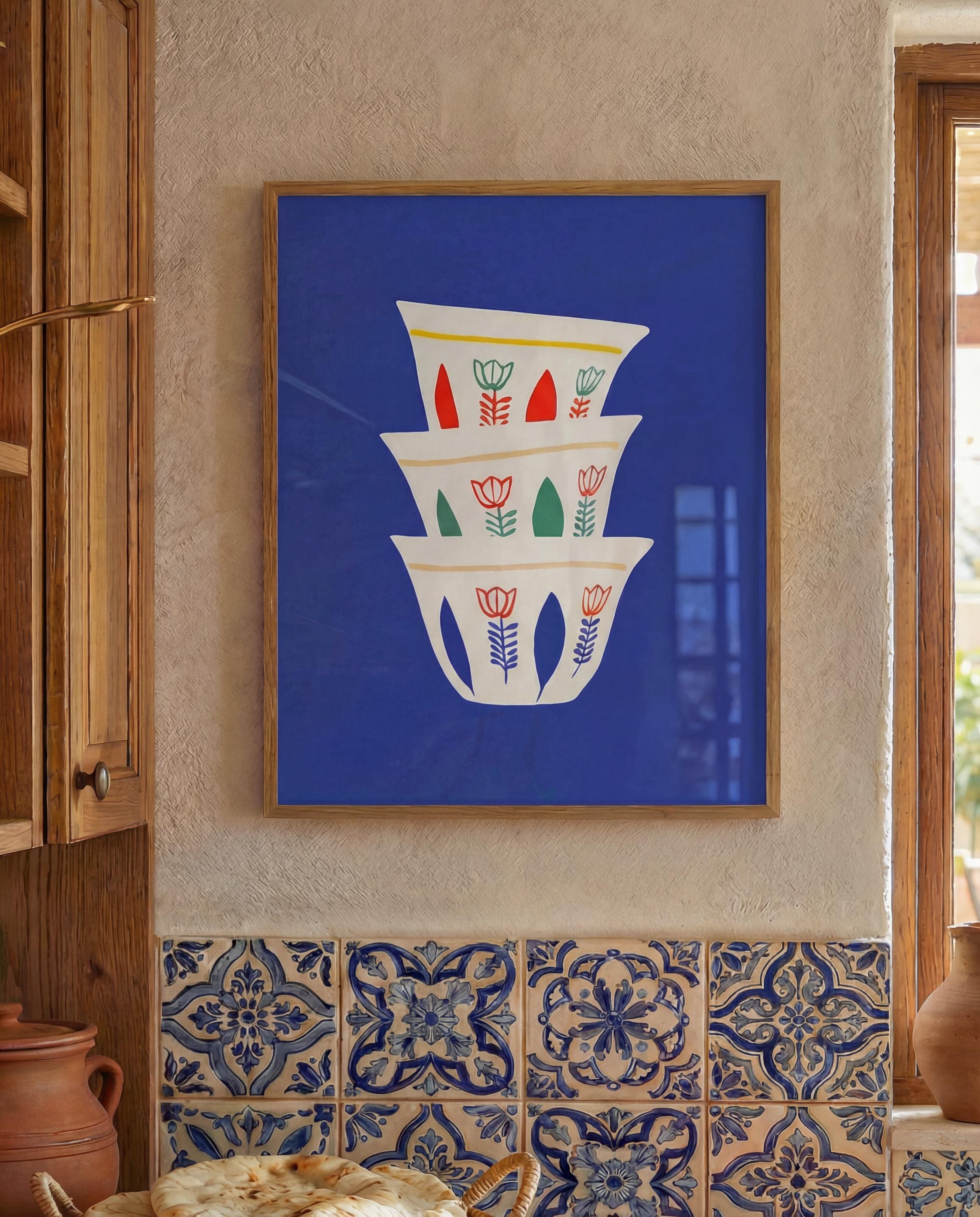 A framed art print featuring a stack of three white coffee cups with floral designs in red, green, and yellow against a vibrant blue background.