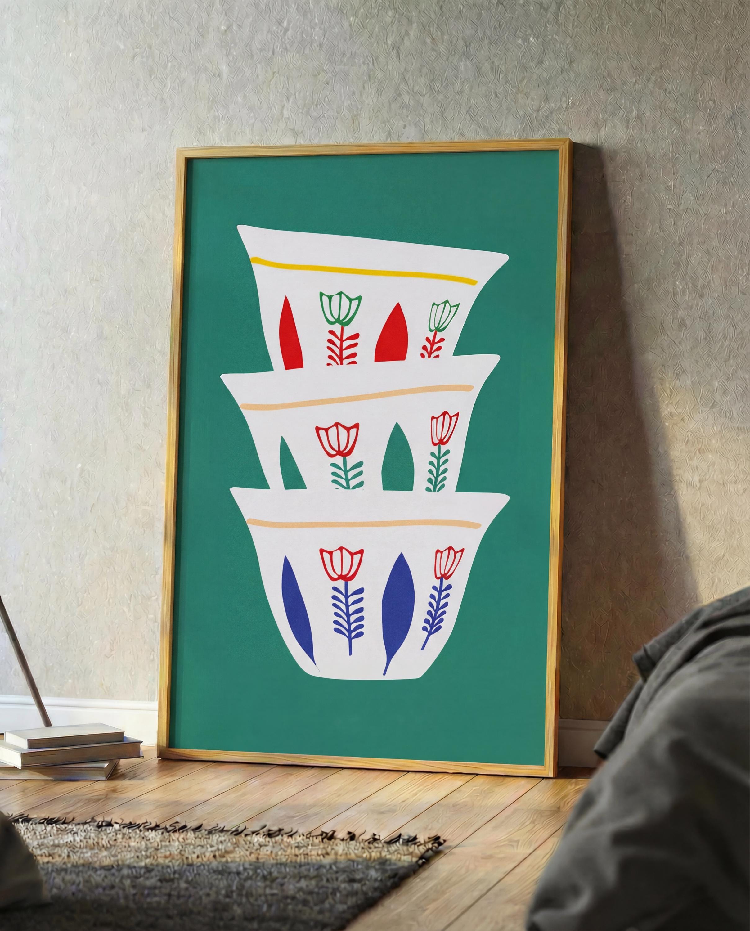 A framed art print featuring a stack of white, stylized coffee cups with floral designs in red, green, and blue against a teal background. The cups have yellow and orange accents. The frame is a light wood color.