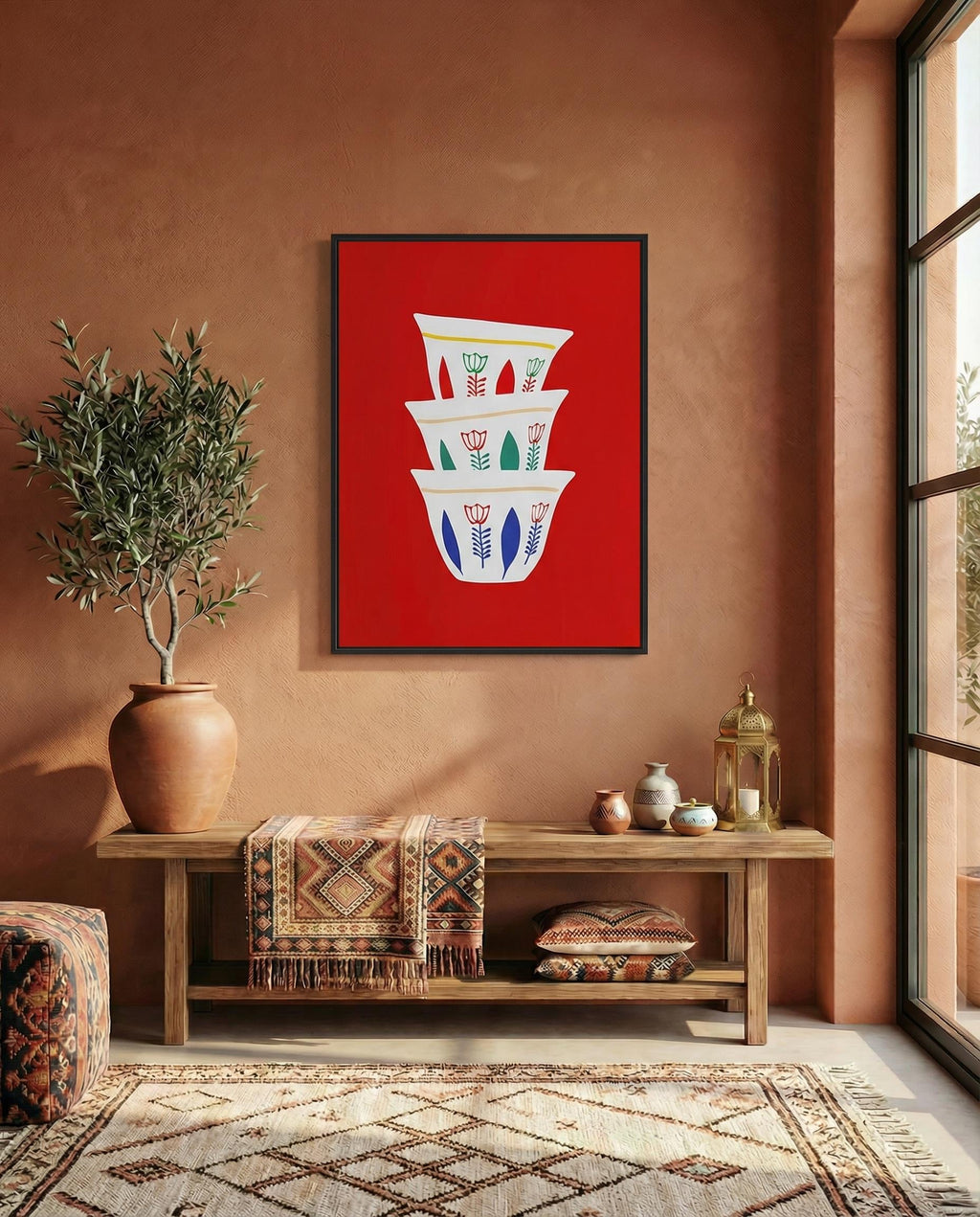 A framed art print featuring a stack of three white coffee cups with floral designs against a vibrant red background. The cups have blue, green, and yellow accents. The artwork is displayed above a wooden bench.