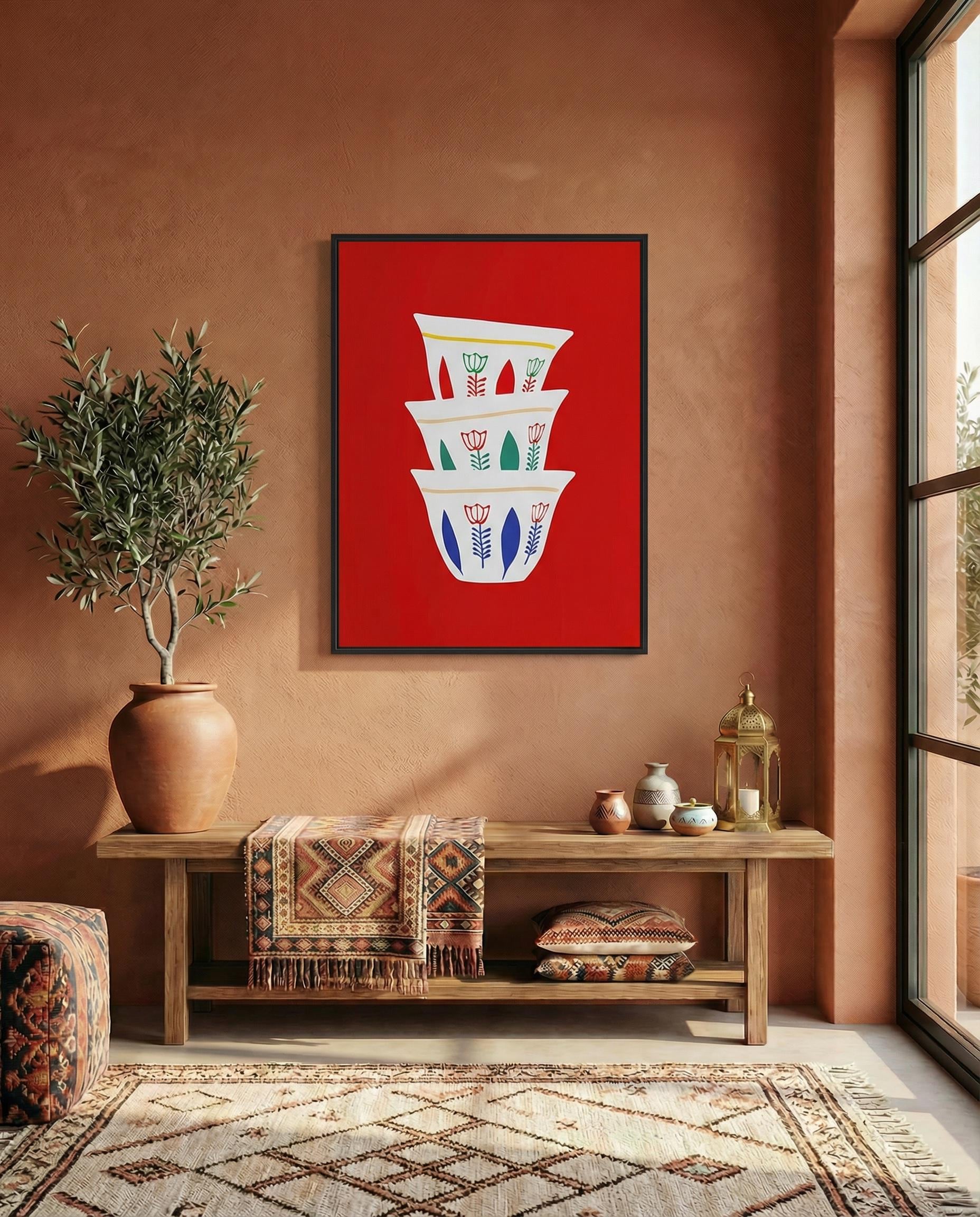 A framed art print featuring a stack of three white coffee cups with floral designs against a vibrant red background. The cups have blue, green, and yellow accents. The artwork is displayed above a wooden bench.